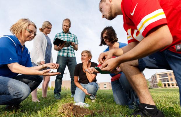 Professor with students looking at dirt samples in a grassy field 
